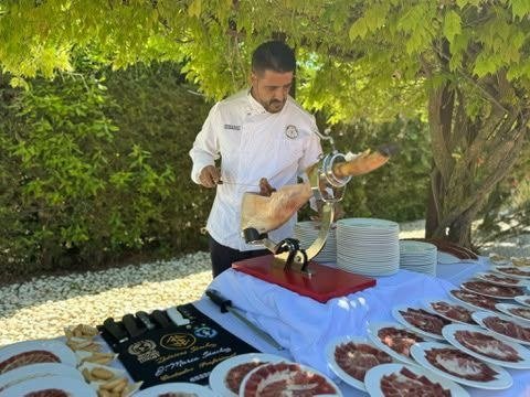 Cortador profesional loncheando una pieza de jamón ibérico en directo ante los invitados, con platos ya servidos en la mesa del evento